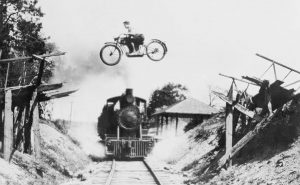 sort hvid billede af ung mand på motorcykel der flyver over en ødelagt bro mens et godstog kører under på skinnerne